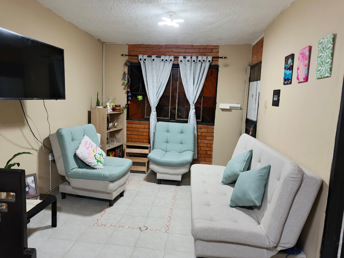 A spacious living area features two light blue chairs and a light gray sofa bed arranged around a neutral rug. Natural light enters through large windows, framed by flowing white curtains. A television is mounted on the wall, and decorative wall art adds a touch of color.