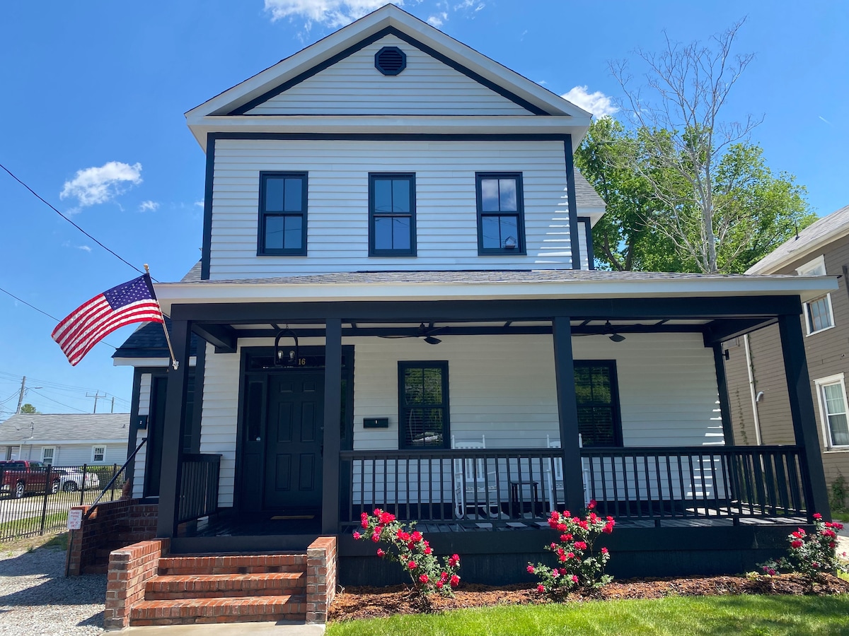 The exterior of the historic Thaddeus C. Dickinson house is showcased, featuring a two-story structure with white siding and black trim. A covered front porch with ceiling fans is visible, framed by blooming rose bushes and an American flag gently waving in the breeze.