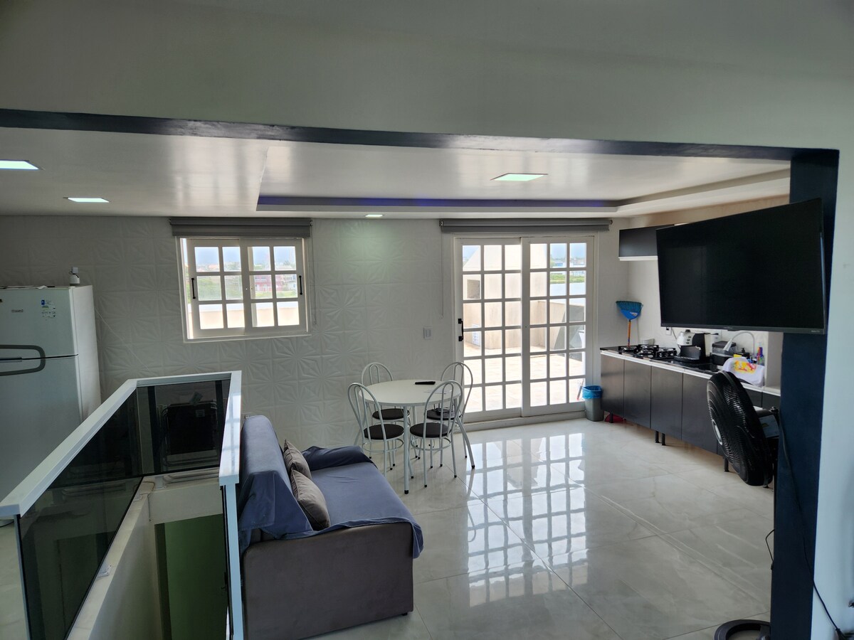 An open living area is shown, featuring a comfortable sofa and a round dining table with four chairs. A large window allows natural light to brighten the space. A television is mounted on the wall, adjacent to a compact kitchen area with modern appliances.