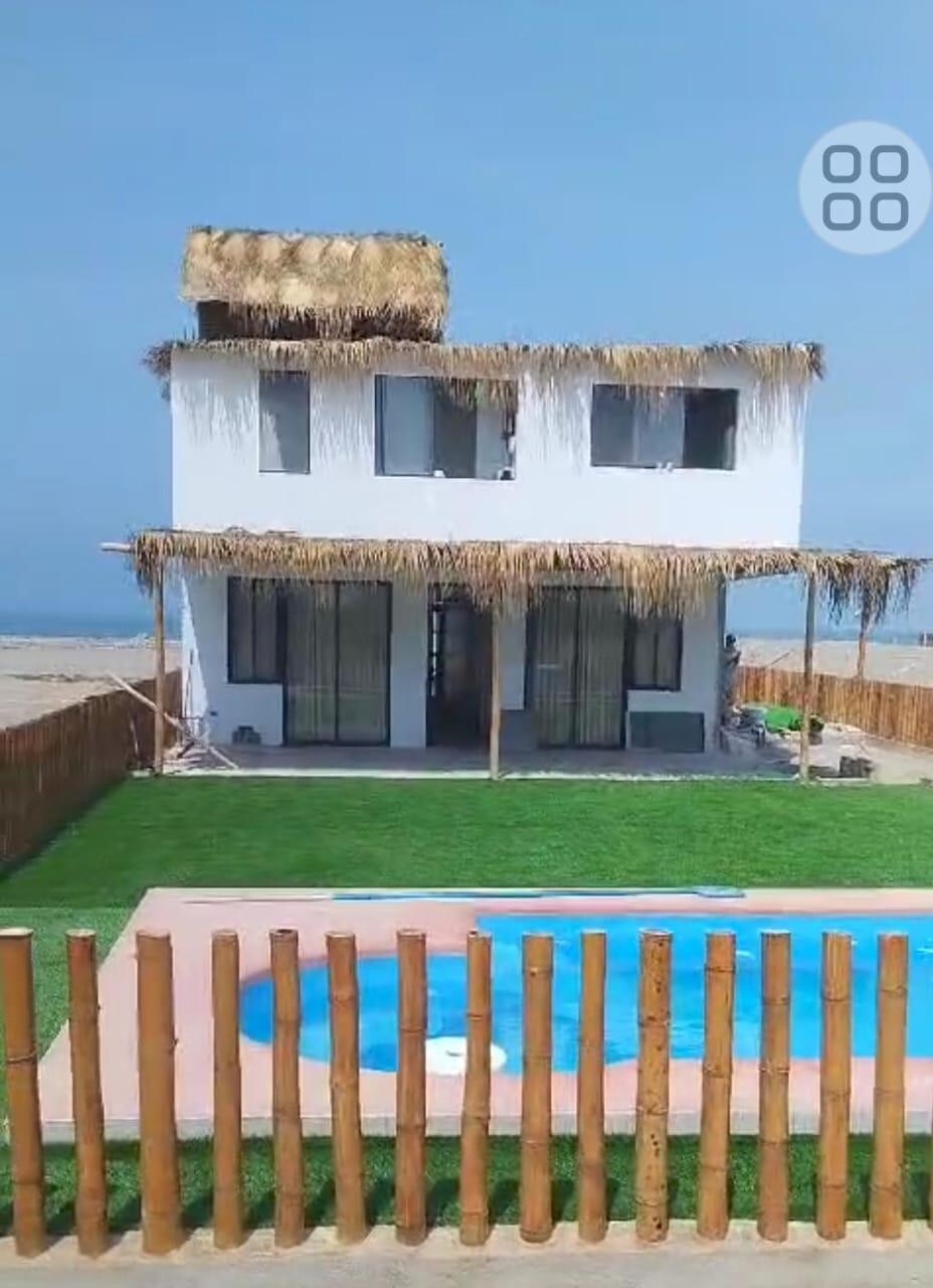 A two-story house with a thatched roof sits near the coast, featuring large glass windows that allow natural light. A manicured lawn and a swimming pool are visible in the foreground, enclosed by a bamboo fence, creating a serene outdoor space.