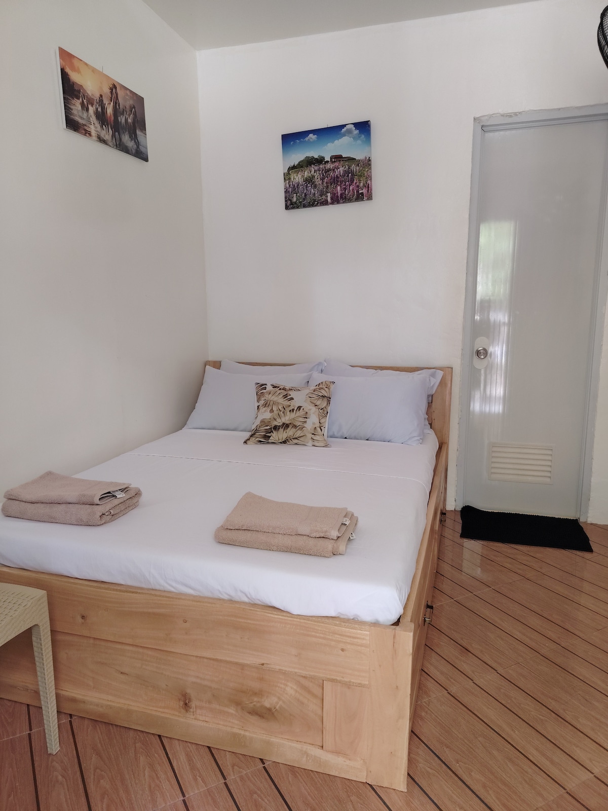 A clean and minimal sleeping area is furnished with a spacious bed dressed in white linens. Two decorative pillows add a touch of style. Light wood accents can be seen in the bed frame and nearby furniture. Artwork is displayed on the walls, enhancing the peaceful ambiance.