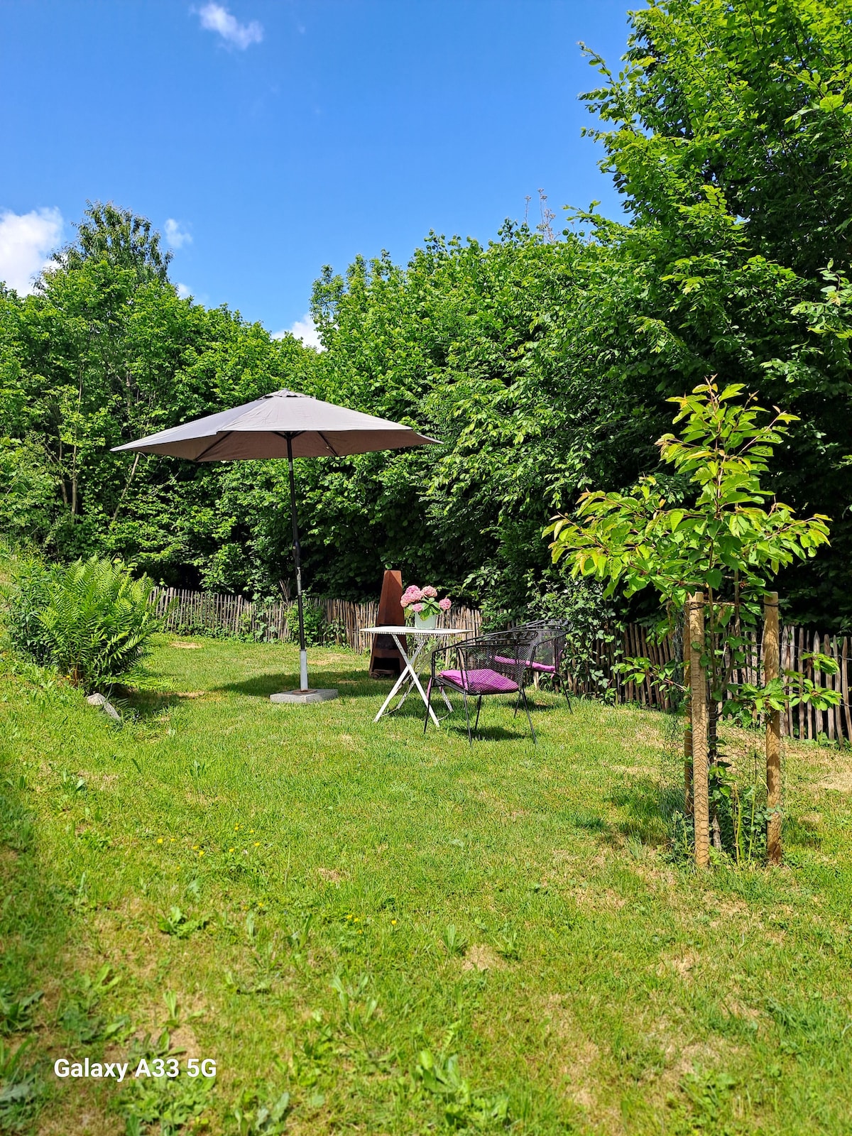 A private garden space features a small table with two chairs beneath an umbrella, surrounded by lush greenery. The area is enclosed by a wooden fence, and a young tree is visible, adding to the tranquil setting.