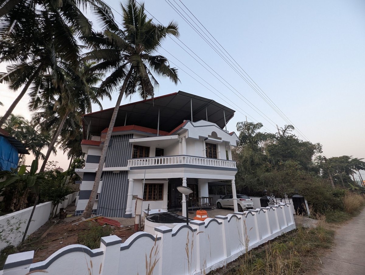 A two-story house is shown, featuring a spacious balcony on the upper level. The exterior is painted in white, complemented by gray accents and surrounded by greenery. A pathway leads to the entrance, with palm trees visible in the background.