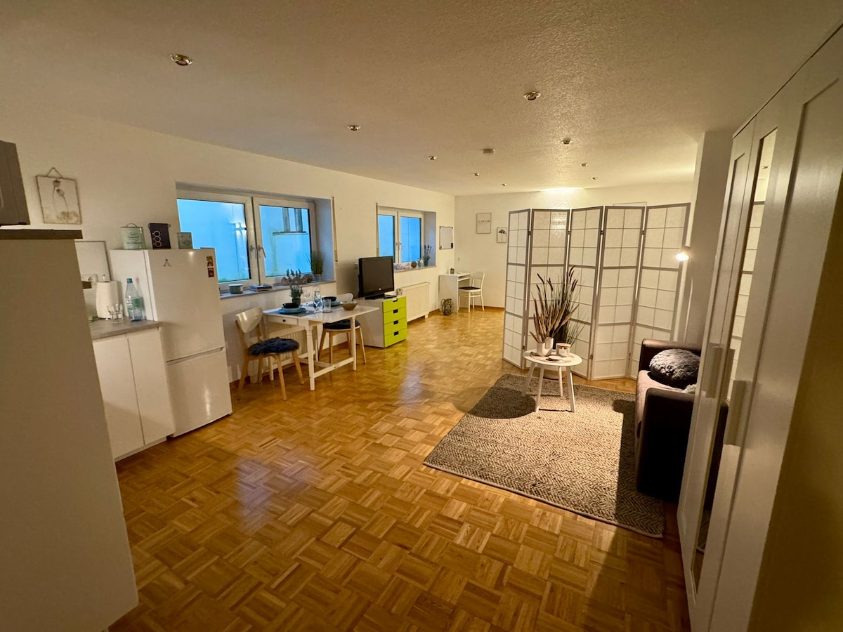 A spacious living area features a light wood parquet floor and is divided by a decorative partition screen. A cozy seating area is present alongside a small table with an arrangement of plants. Natural light floods in through large windows, illuminating the modern decor.
