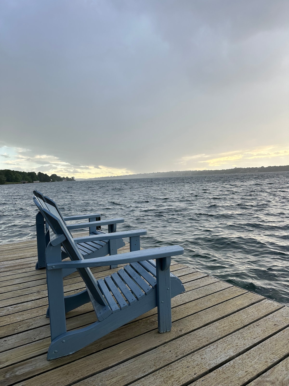 Two blue Adirondack chairs are positioned on a wooden dock extending over calm water. The surface of the water reflects a soft light as the sky transitions from cloudy to a subtle glow, creating a serene and inviting view.