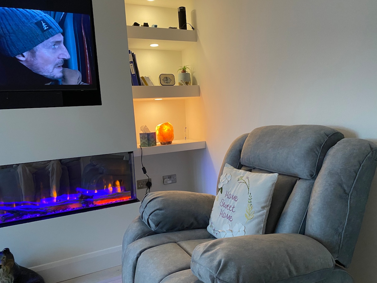A comfortable recliner is positioned near a modern electric fireplace, which is illuminated in soft blue and orange hues. Above, a wall-mounted television is visible. Floating shelves display decorative items, including a glowing salt lamp, creating a cozy ambiance.