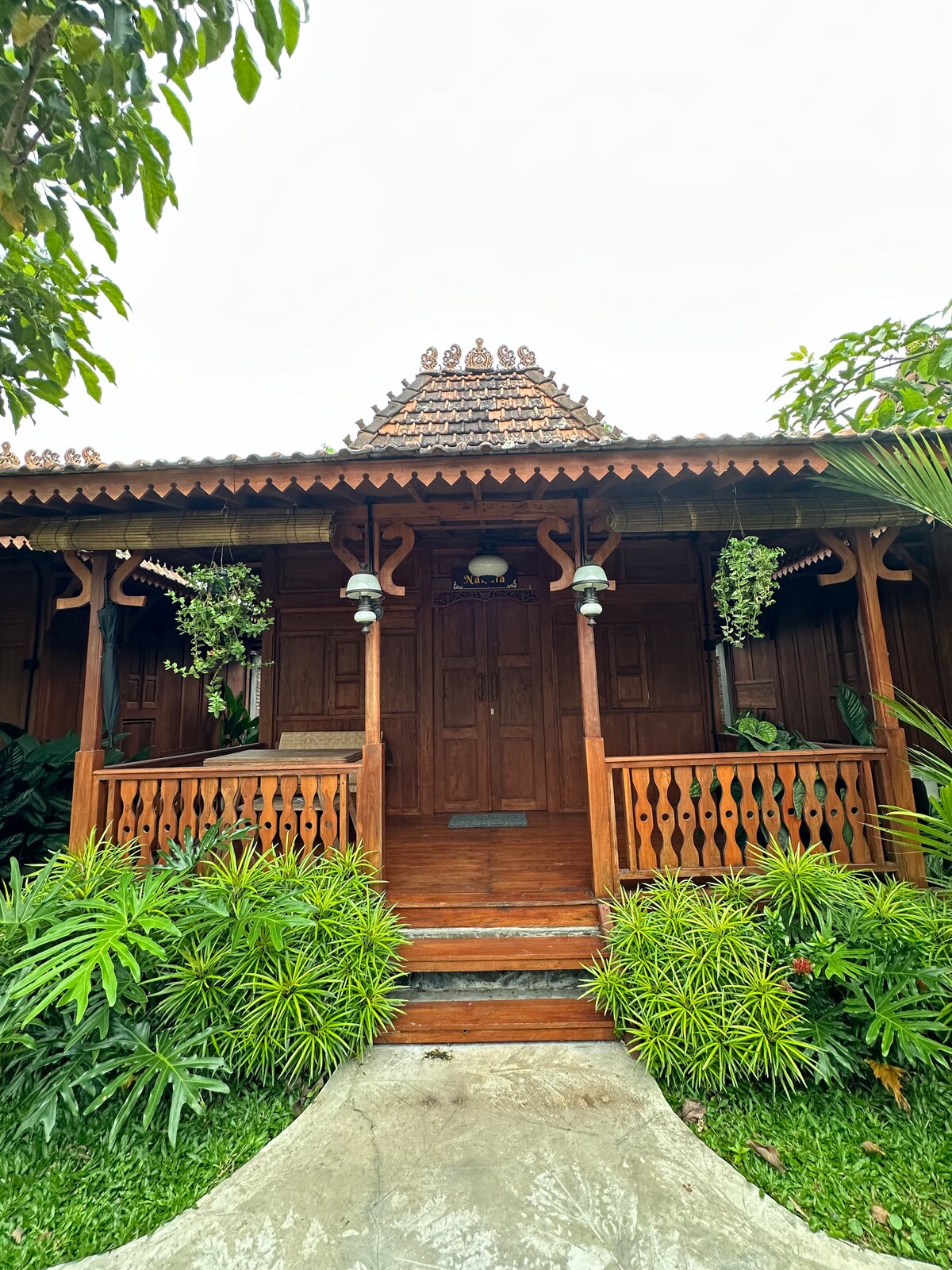 The entrance of a wooden cottage is framed by lush greenery, featuring a decorative roof and traditional carvings. Two pillars support the porch, adorned with hanging plants. A clear path leads to the wooden double doors, inviting guests into the peaceful setting.
