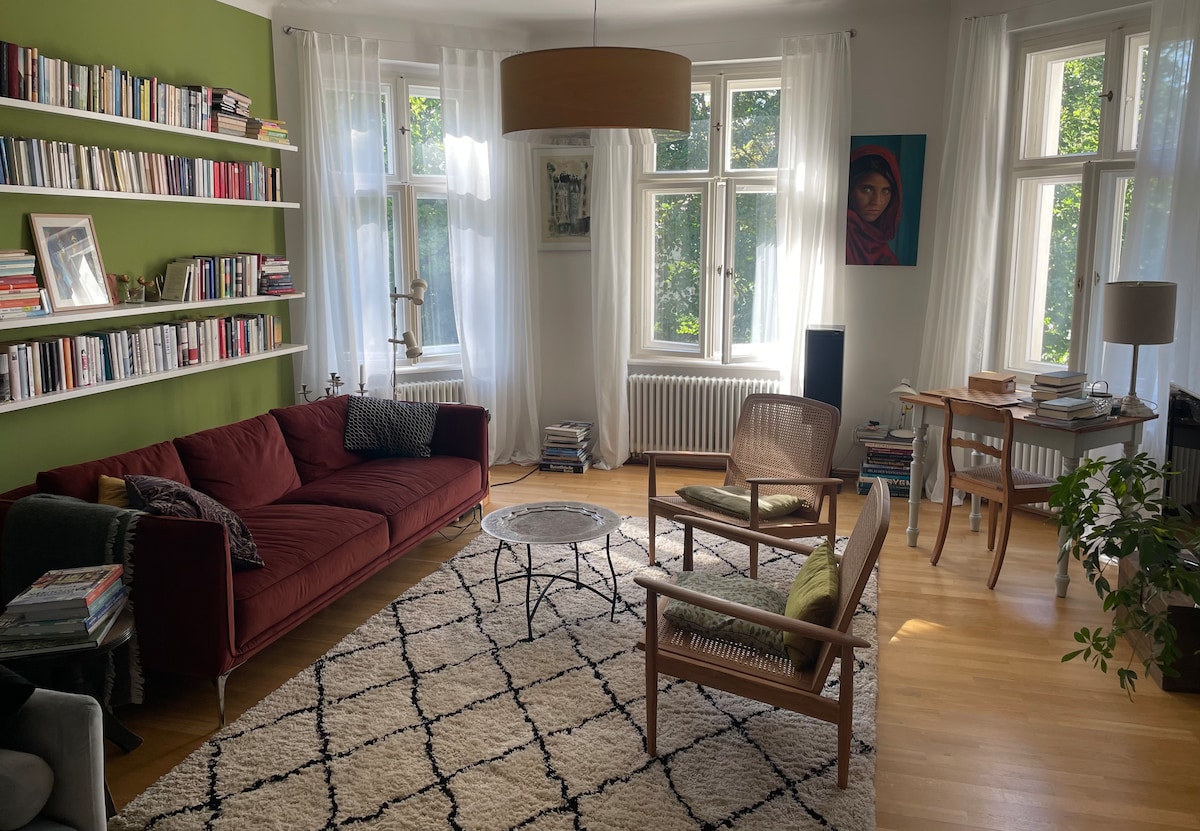 A living area showcases a deep red sofa, framed by a wall of bookshelves. Natural light floods through large windows, illuminating a circular coffee table positioned on a patterned area rug. A workspace is visible, featuring a desk and chair next to an indoor plant.