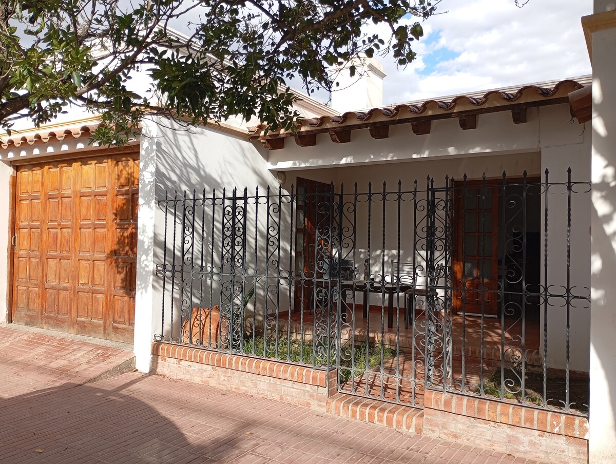 A front view of the house features a rustic wooden garage door and a decorative wrought iron fence. The spacious porch is visible, showcasing a seating area with a table. Light filters through the trees, creating playful shadows on the brick pathway.
