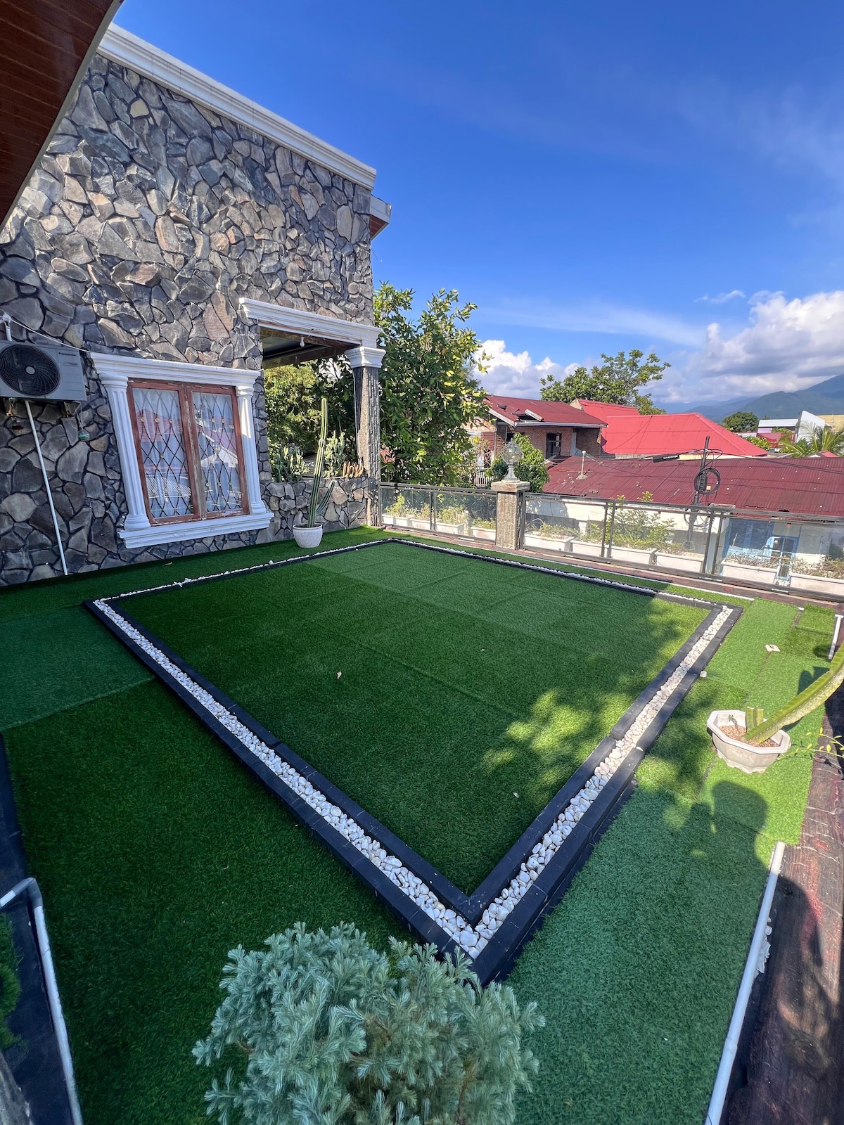 A spacious outdoor area is highlighted by well-maintained artificial grass surrounded by decorative stones. The stone exterior of the building is visible, along with a clear blue sky and nearby rooftops framed by greenery.