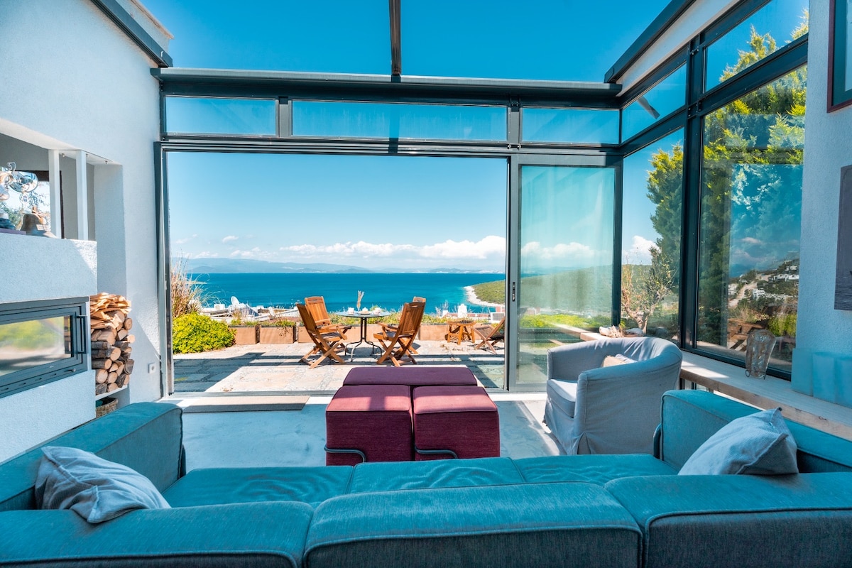 A modern living area features large glass doors that open to a stunning sea view, blending indoor and outdoor living. Comfortable seating arrangements include a spacious sofa, two armchairs, and a coffee table. Natural light fills the space, enhancing the serene ambiance.