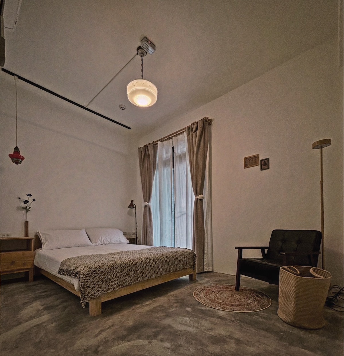 A serene bedroom features a spacious bed with a patterned coverlet, complemented by a simple headboard. Natural light filters through sheer curtains covering a large window. A cozy chair and a woven basket add functionality and warmth to the minimalist decor.