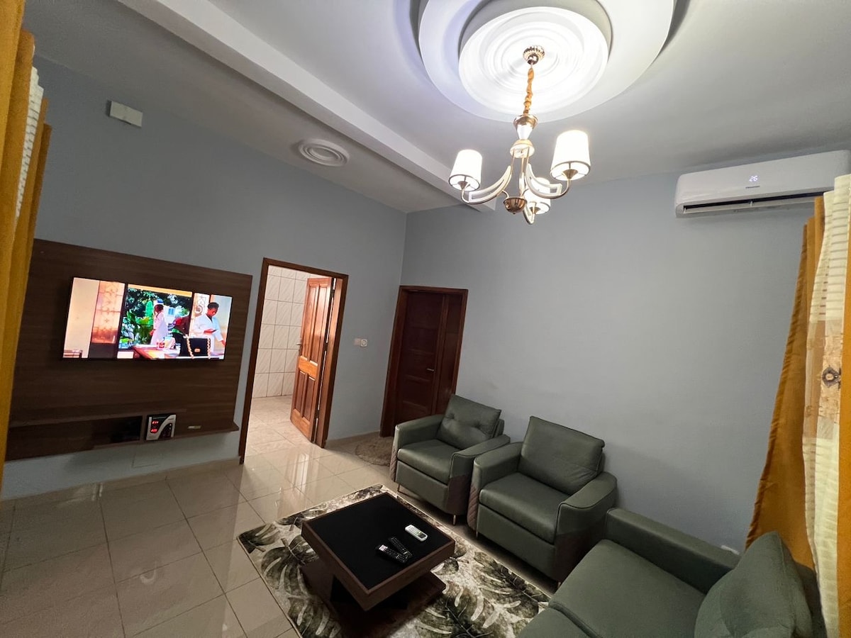 A living area features a modern design, with two armchairs and a coffee table situated on a patterned rug. A flat-screen television is mounted on the wall. Natural light enters through the window, which is adorned with light yellow curtains. A decorative chandelier hangs from the ceiling.