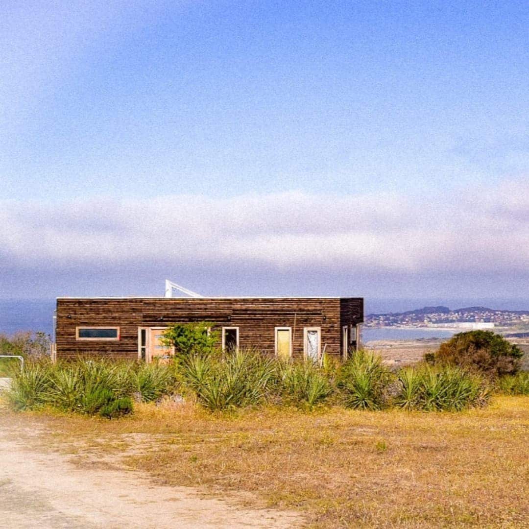 A modern wooden house is set against an open landscape, surrounded by native greenery. The structure features large windows, providing views of the horizon and distant coastal scenery. A clear sky hints at the natural light enjoyed inside, enhancing the connection to the tranquil environment.