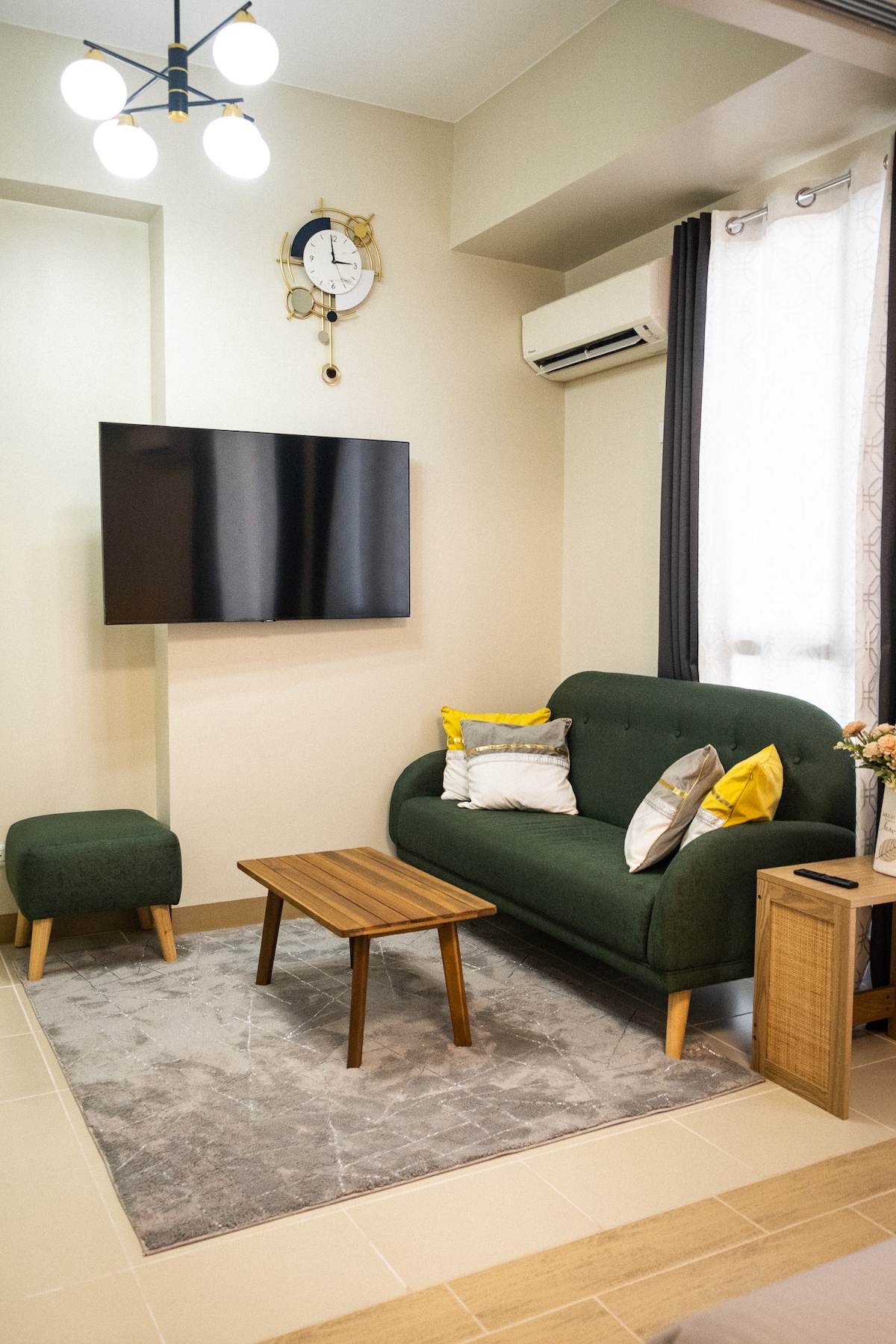 A stylish living area is presented with a green two-seater sofa and a matching ottoman. A wooden coffee table sits on a soft area rug, while a wall-mounted TV is visible. Natural light enters through sheer curtains, complemented by a decorative clock above.