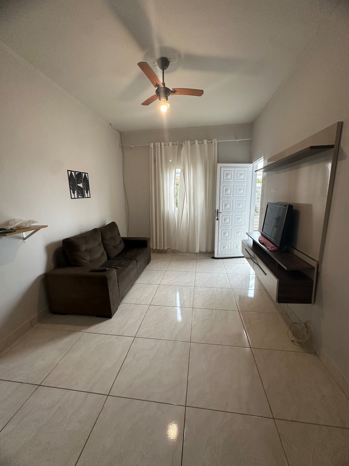 A well-lit living area is presented with a comfortable brown sofa and a wall-mounted television. Natural light enters through a window covered with sheer curtains, offering a soft ambiance. The glossy floor tiles enhance the brightness of the space.