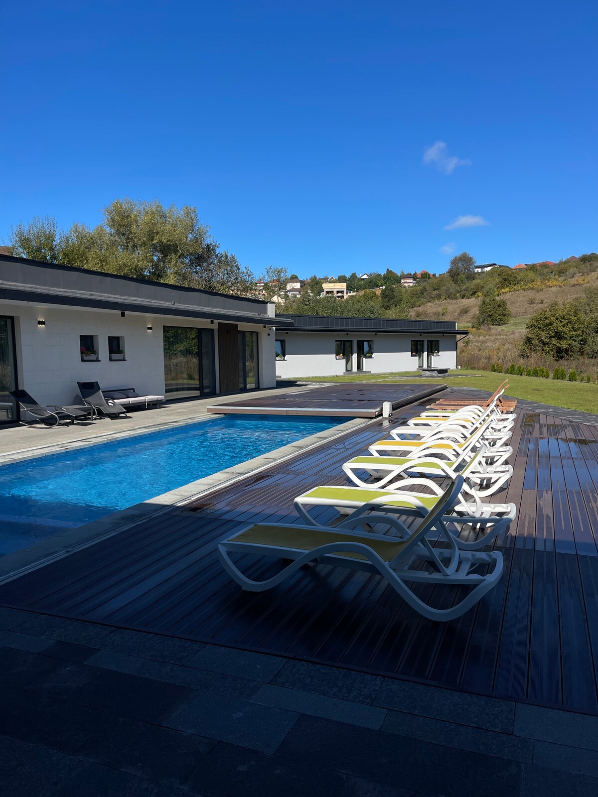An outdoor area is seen with an inviting swimming pool surrounded by multiple lounge chairs. A separate hot tub is located beside the pool, and the landscape features greenery and a hillside in the background, complementing the serene environment.