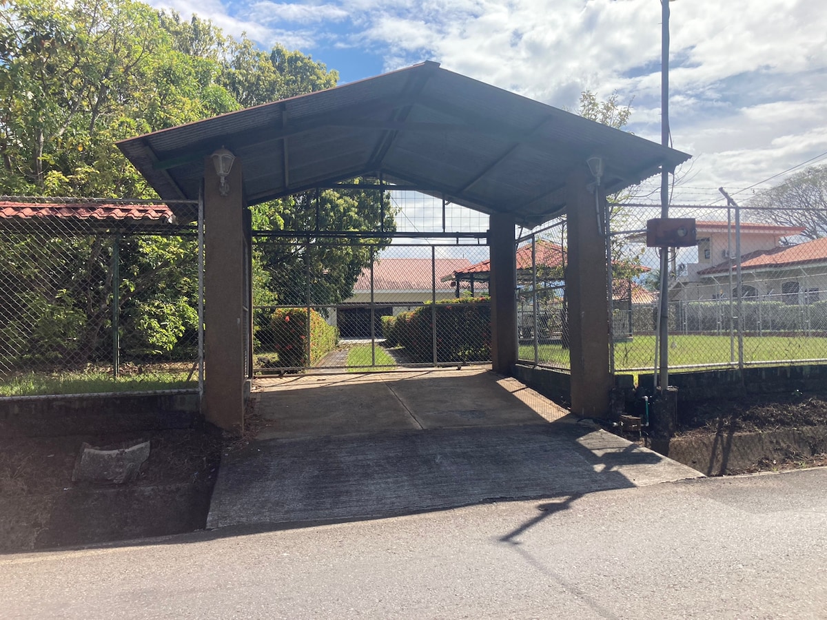 The entrance features a covered gate providing access to the property. A well-maintained fence surrounds the area, complemented by lush greenery in the background. The driveway is paved, leading into a spacious area beyond the gate.