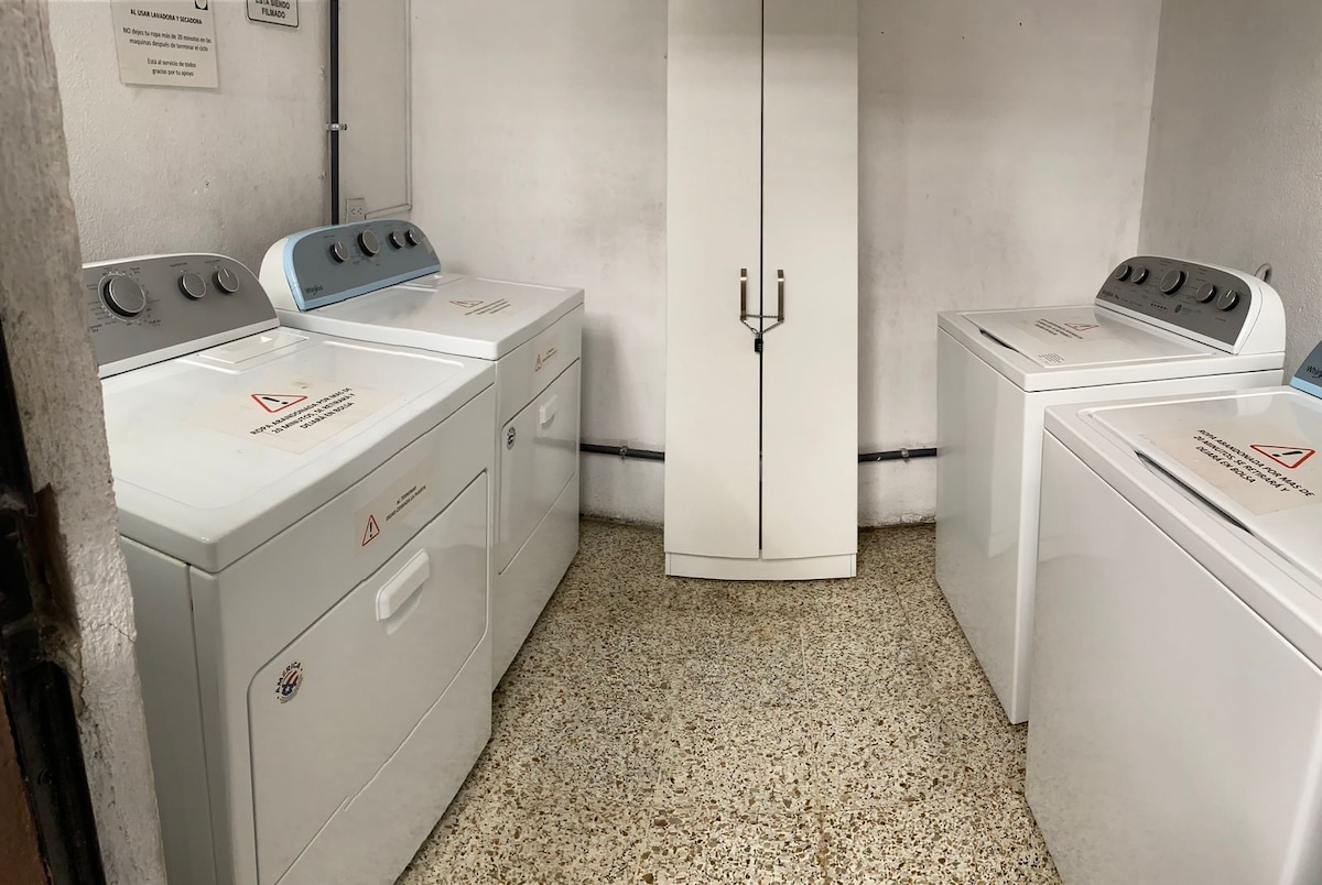 A laundry area features three washing machines and a dryer, each equipped with controls at the top. A tall cabinet with handles is positioned in the corner, and the flooring is composed of light-colored speckled tiles.