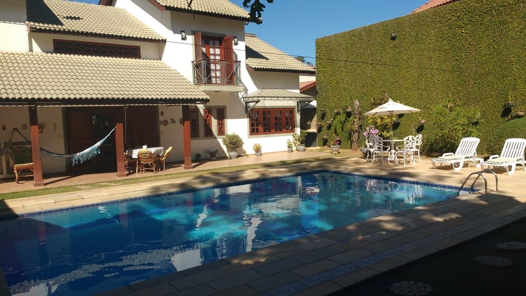 A spacious outdoor area features a clear swimming pool surrounded by sun loungers and an umbrella-shaded dining set. A hammock is seen strung between two posts, and lush greenery provides privacy. The charming house displays a Mediterranean-style tiled roof and multiple windows.