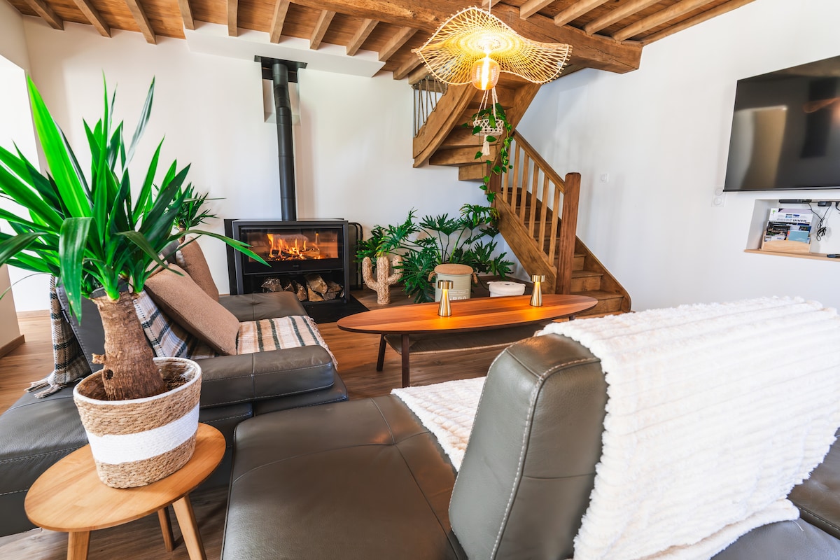 The living area features a blend of comfortable seating and greenery, highlighted by a central fireplace. Wooden beams adorn the ceiling while a staircase leads to the upper floor. Soft textiles and natural elements create an inviting space for relaxation.