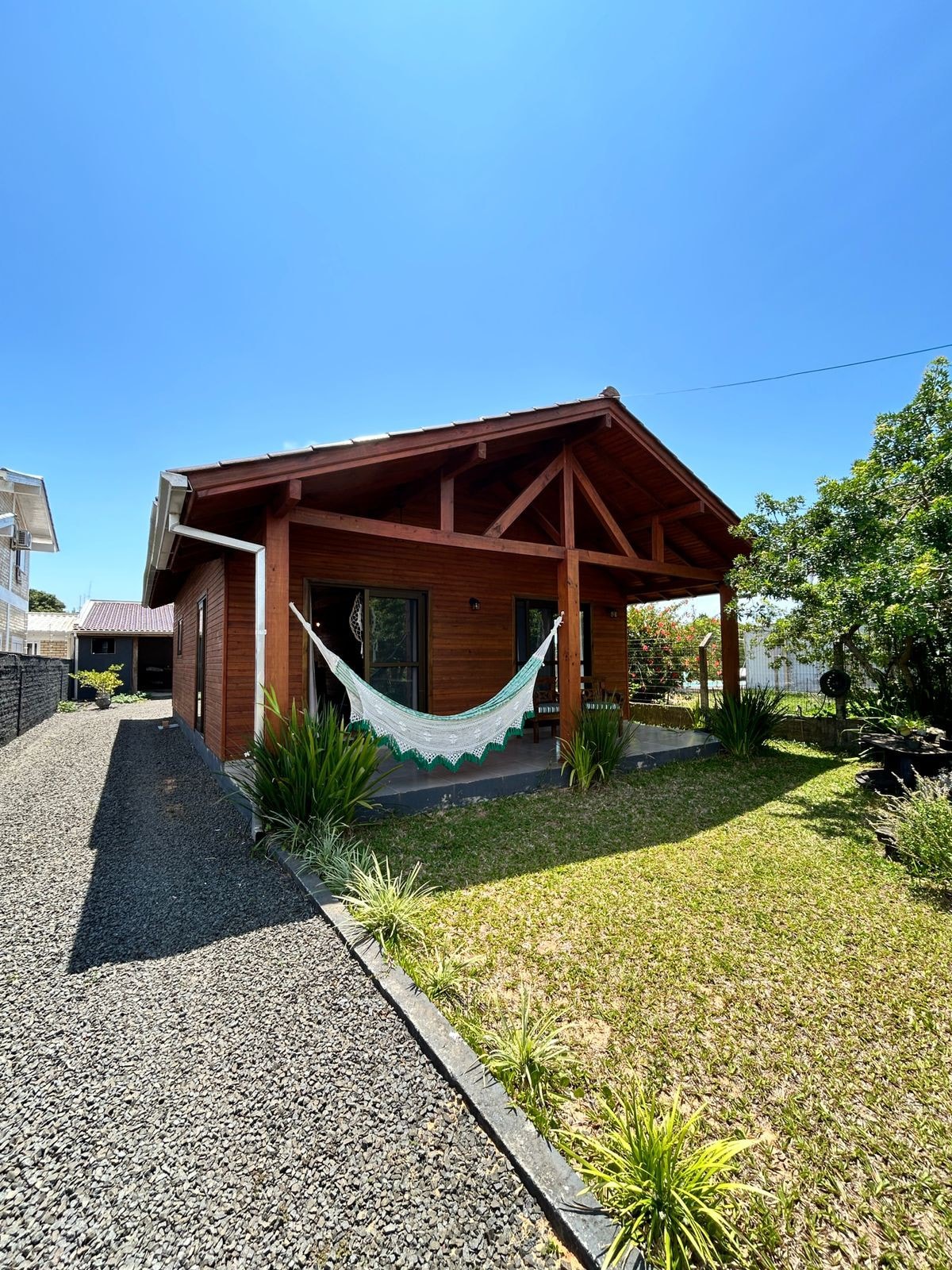 A charming wooden bungalow is featured, set against a clear blue sky. A welcoming hammock is hung on the porch, surrounded by lush greenery and well-maintained grass. The gravel path leads to the entrance, enhancing the overall tranquil vibe of the space.