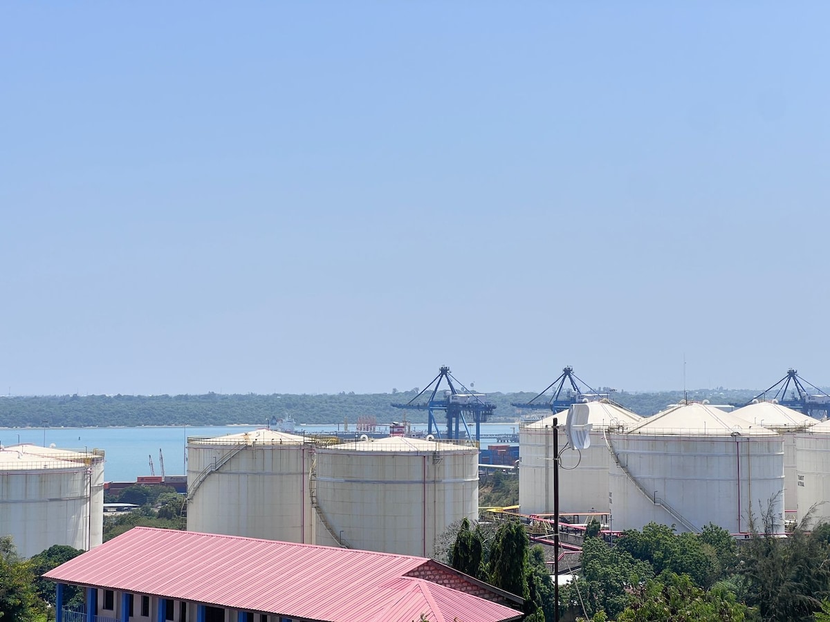 Large storage tanks stand prominently in the foreground, with a vibrant blue sky above. A calm body of water and distant structures are visible in the background, suggesting a port area. Lush greenery can be seen surrounding the base of the tanks.