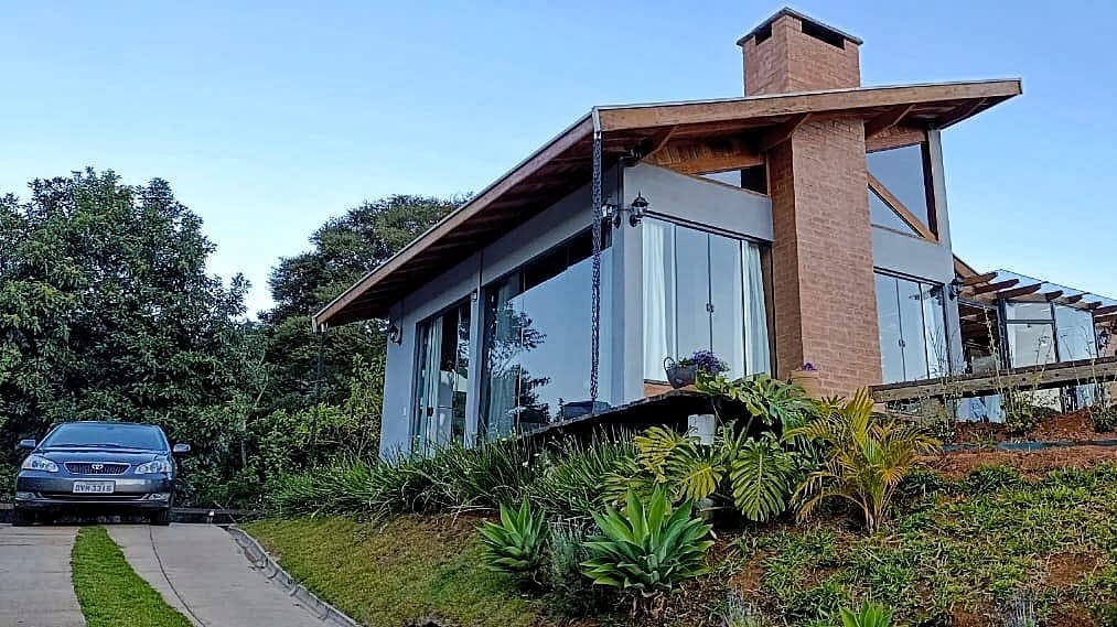 The exterior of the chalé showcases a modern design with large glass windows reflecting its natural surroundings. A sloped driveway leads to a private garage, while surrounding greenery and a stone pathway enhance the connection to the landscape. A chimney adds a rustic touch.