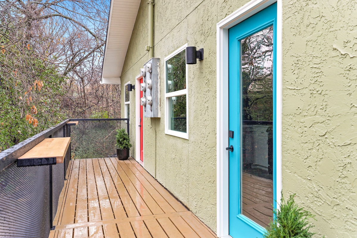 A wooden deck is visible, featuring a smooth surface with a railing that provides a view of the surrounding trees. Two colorful doors, one red and one turquoise, are positioned against a light green exterior wall, enhancing the inviting atmosphere of the space.