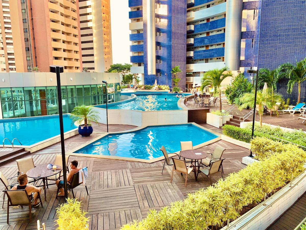 An inviting outdoor pool area features multiple swimming pools surrounded by sun loungers and tropical plants. Wooden decking provides space for seating arrangements with tables and chairs, creating a relaxing atmosphere. High-rise buildings rise majestically in the background, highlighting the urban setting.