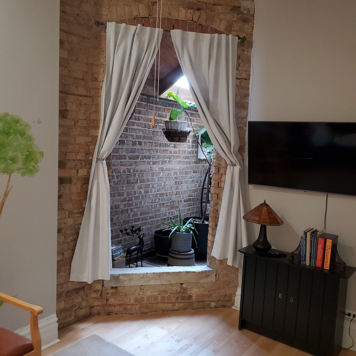 An open window is framed by white curtains that draw back to reveal a small outdoor space filled with potted plants. A brick wall provides a rustic backdrop, while a television mounted on the adjacent wall and a dark cabinet with books sit nearby.