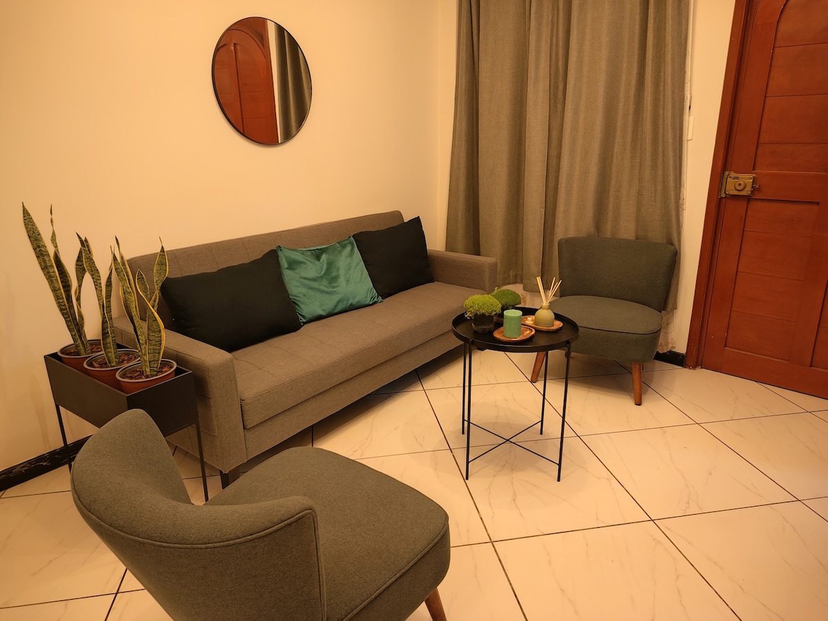 A cozy seating area is centered around a circular table, featuring two gray armchairs and a compact sofa adorned with green and black cushions. Potted plants are arranged near the wall, and a round mirror reflects the soft light in the room.