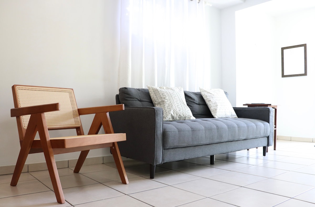 A comfortable living area is shown with a grey couch and two decorative pillows. A wooden chair with a woven backrest complements the space. Natural light is filtered through sheer white curtains, illuminating the tiled floor and a single framed mirror is positioned on the wall.