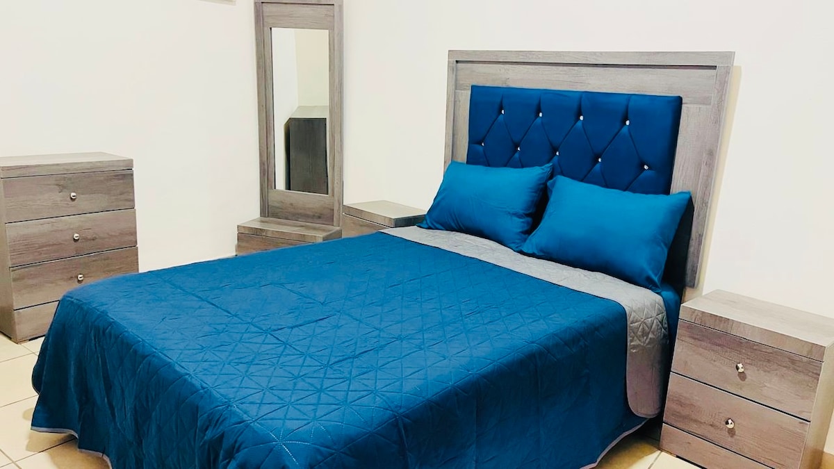 A serene bedroom is furnished with a spacious bed adorned with a blue quilt and accented by two matching pillows. Flanking the bed are two light-colored bedside tables, and a tall mirror complements the decor against a neutral wall.