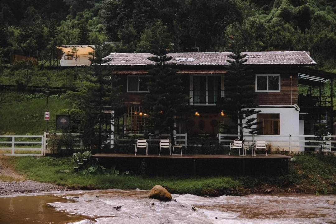A rustic retreat is situated beside a flowing river, surrounded by lush greenery. The two-story building features a wooden exterior with large windows, accompanied by a spacious deck outfitted with several white chairs for outdoor relaxation.