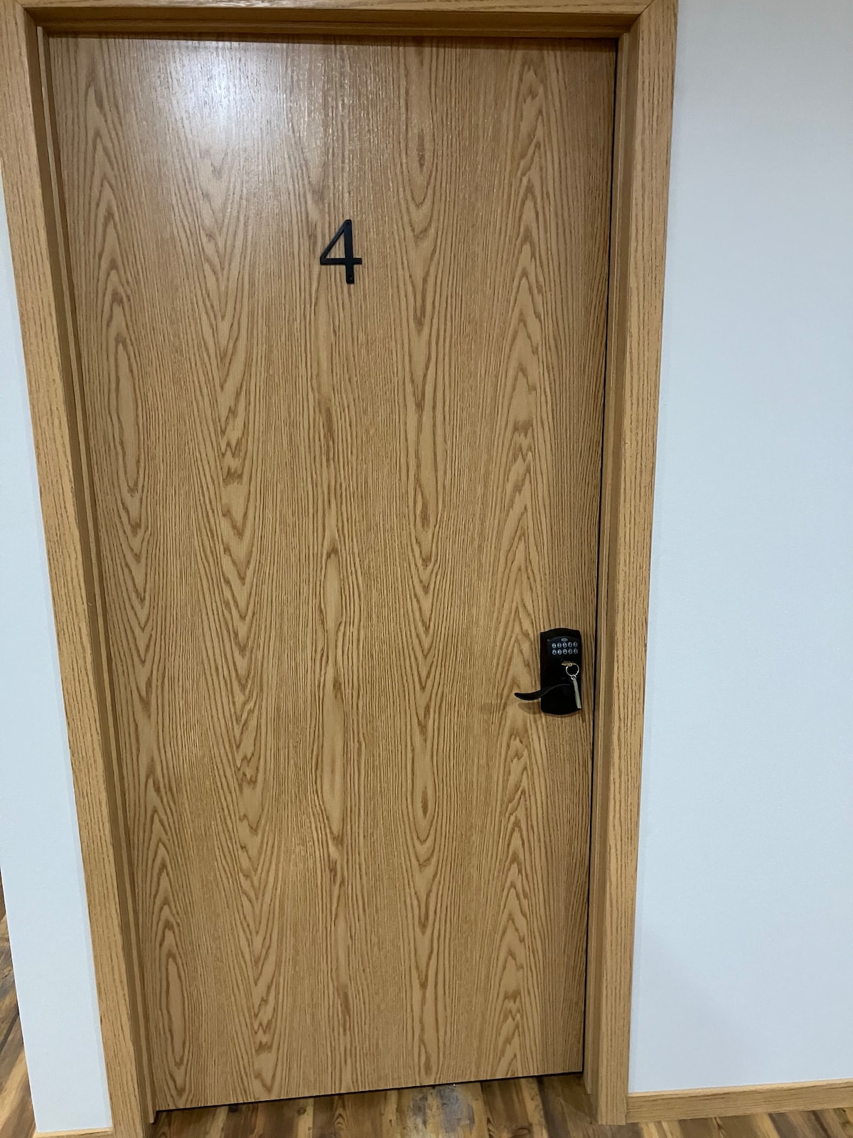A wooden door with a natural finish is shown, featuring the number '4' displayed prominently. A modern handle and lock system are installed, adding a contemporary touch. The wall surrounding the door is painted in a neutral tone, complementing the overall design.