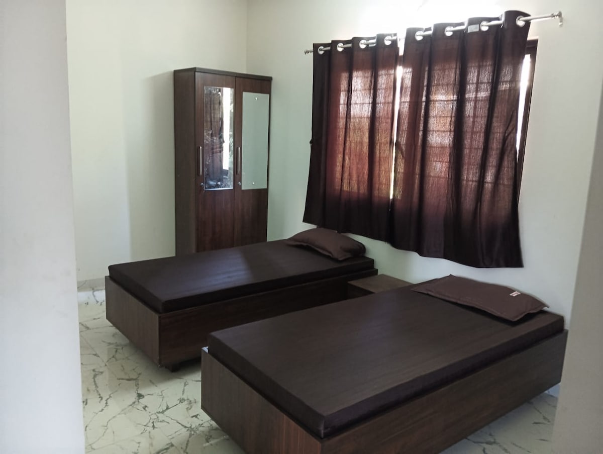 The room features two individual beds with dark wooden frames positioned beside a small nightstand. A tall, mirrored wardrobe reflects natural light, while the floor displays a marbled pattern. Black curtains adorn the window, providing a sense of privacy.