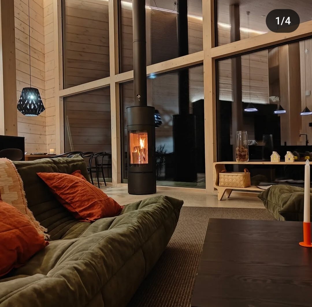 A spacious living area is highlighted by large windows, allowing natural light to fill the space. A modern fireplace stands centrally, surrounded by comfortable seating with decorative pillows. Minimalist decor is present, with a soft area rug underfoot and an elegant dining table visible in the background.