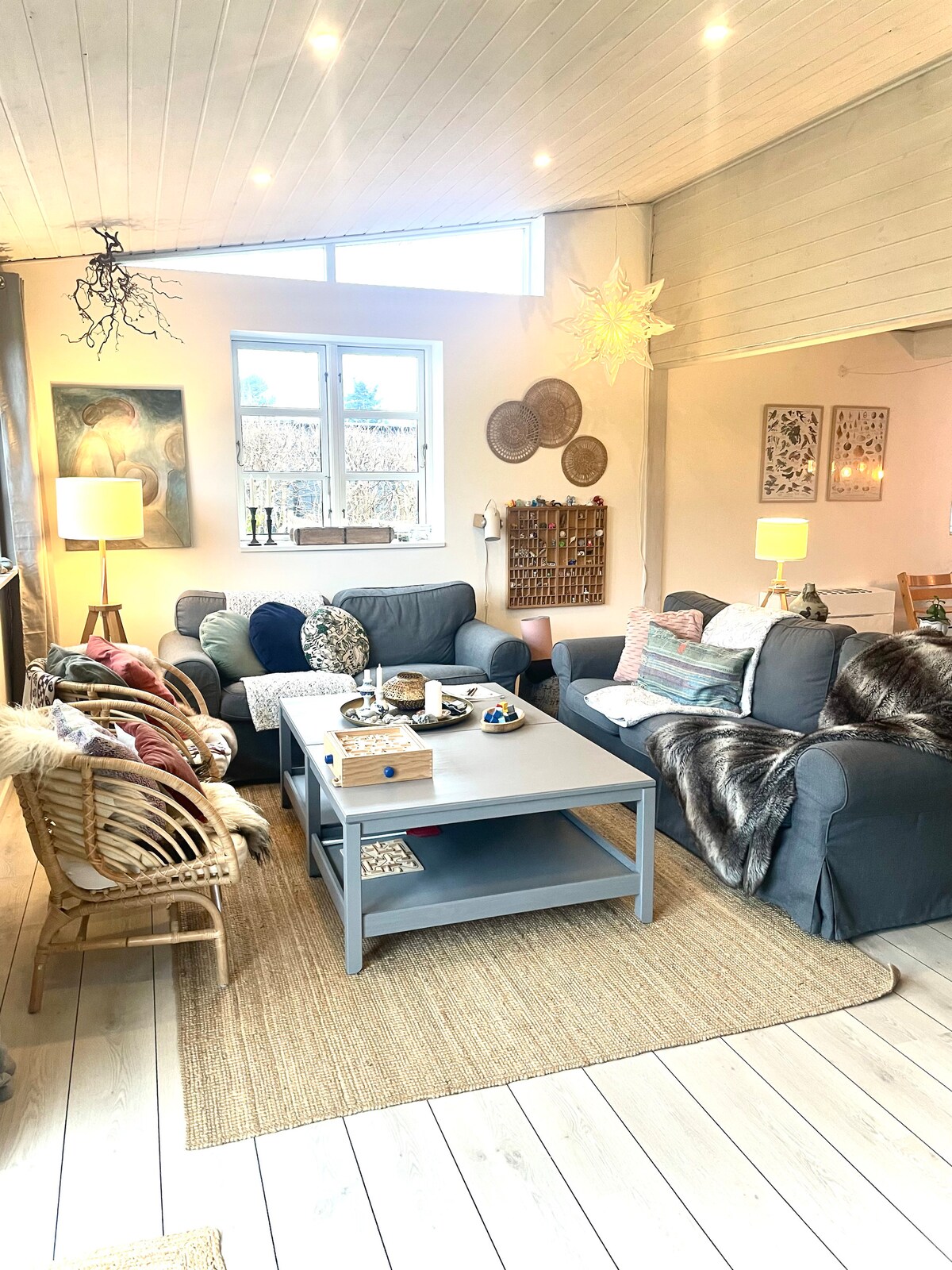 The living area features a combination of comfortable seating, including sofas and a wicker chair. A coffee table is centered among the chairs, adorned with decorative items. Natural light enters through a large window, illuminating the room's cozy decor and inviting ambiance.