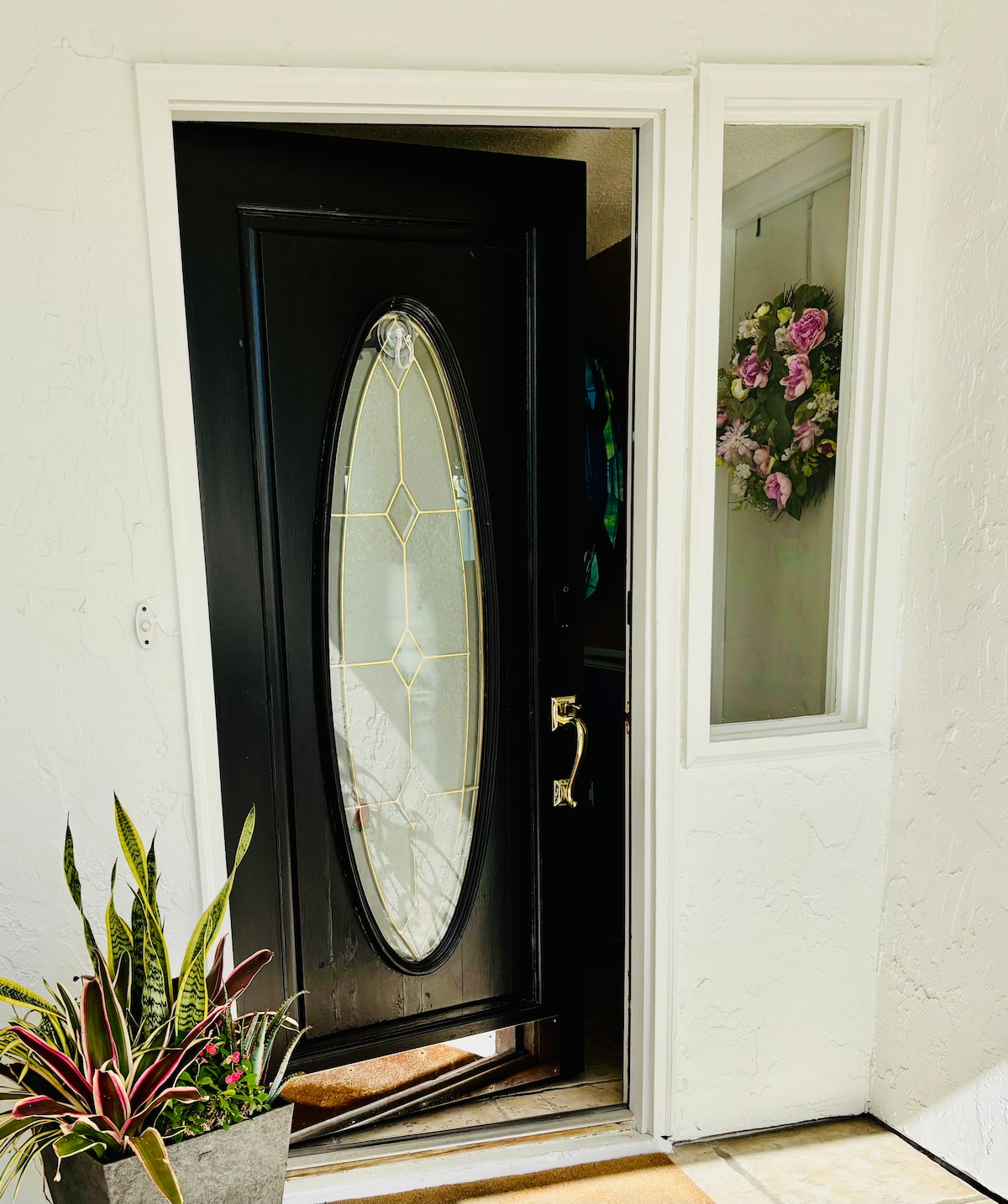 A welcoming door entry showcases a stylish black door with an oval window design, partially opened to reveal inside. A decorative wreath with purple flowers adorns the adjacent wall, while a potted plant adds a touch of greenery to the entryway.