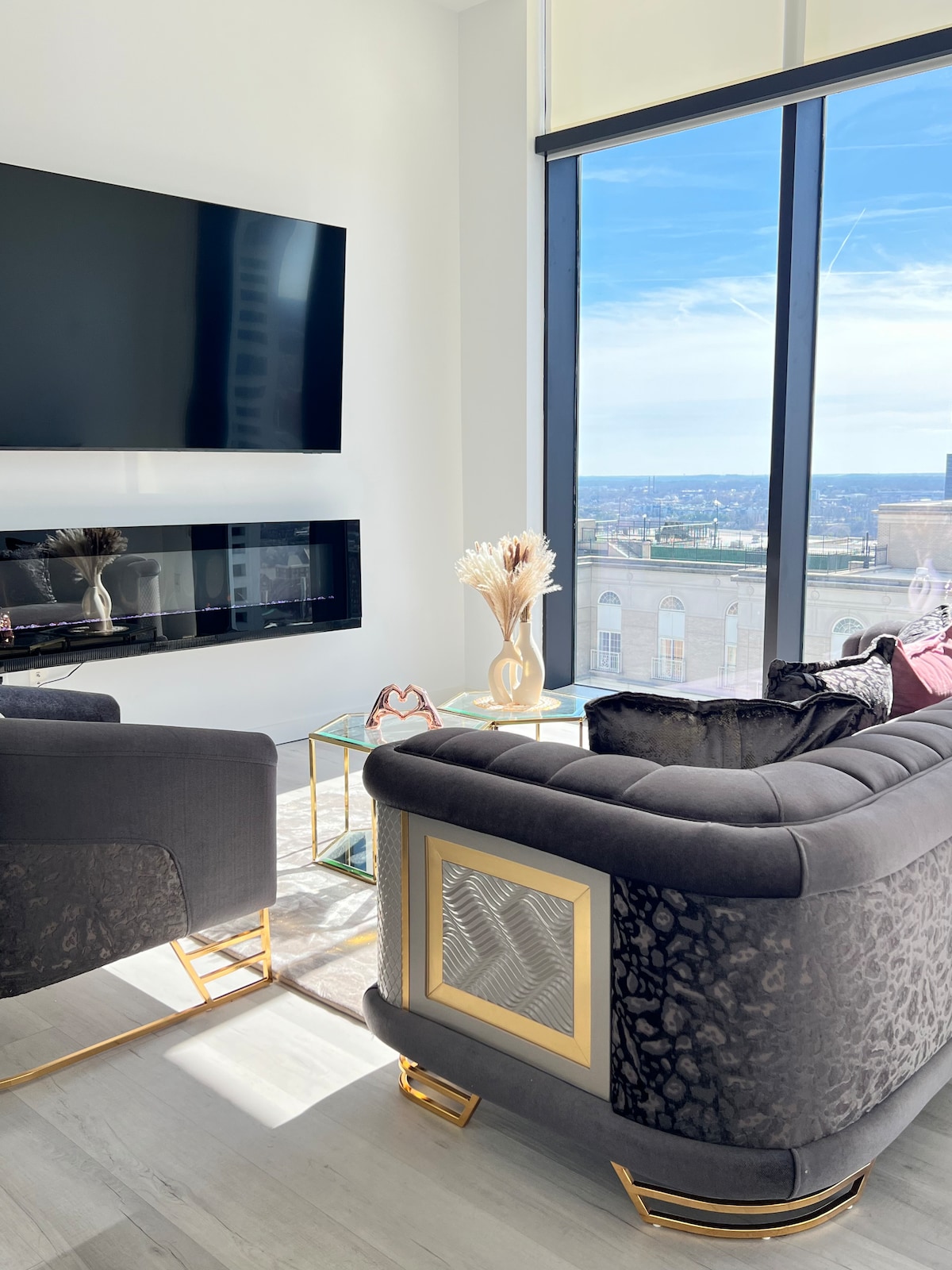 The living area features plush seating with a reflective gold base, positioned near large windows offering expansive views of the cityscape. A modern television is mounted on the wall above an elegant electric fireplace, with a glass coffee table in the center completing the setup.