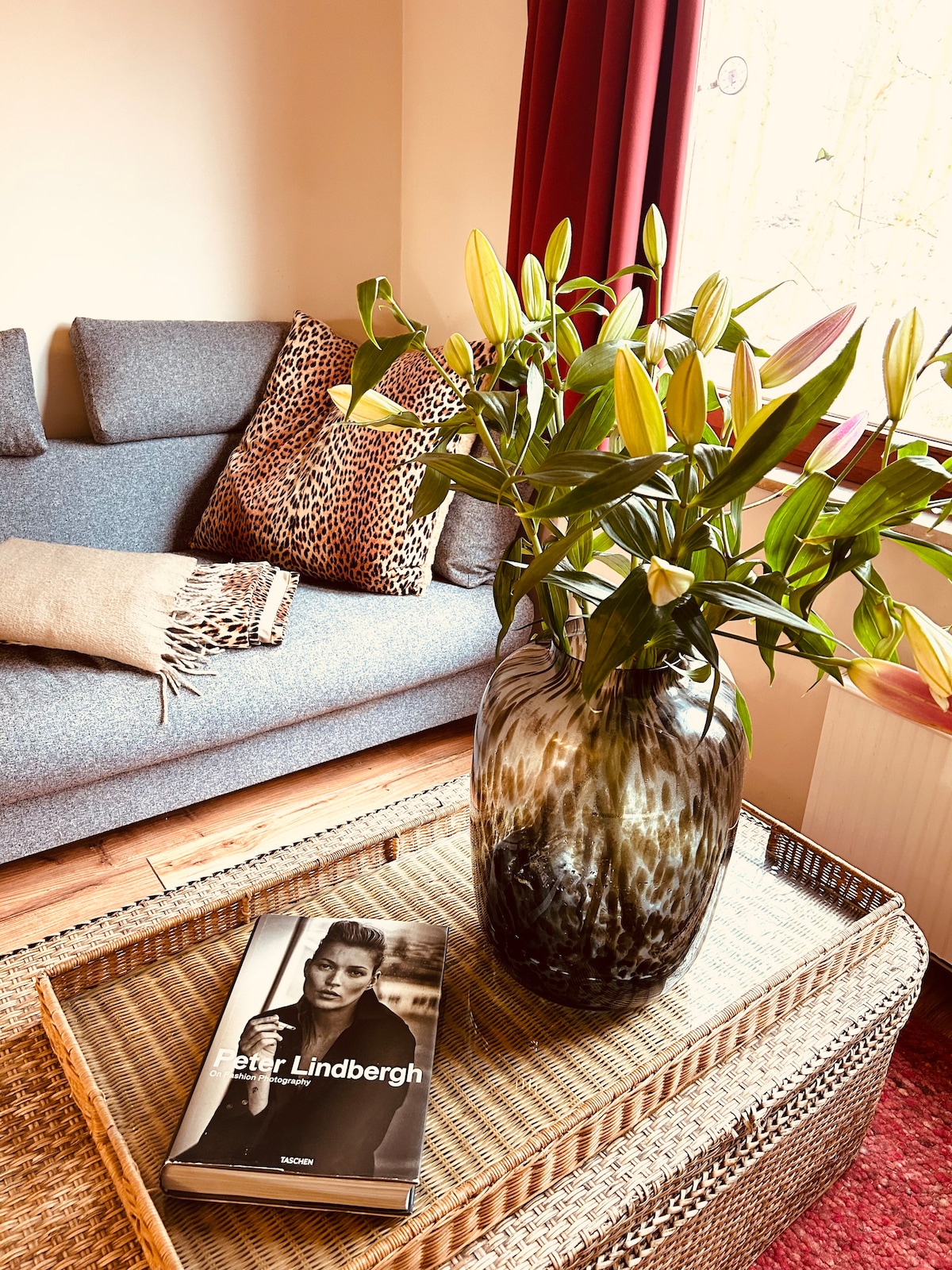 A comfortable seating area features a gray sofa adorned with a textured throw and animal-print cushions. A large vase with fresh lilies sits on a woven coffee table, accompanied by a book on photography, adding a touch of elegance to the space.