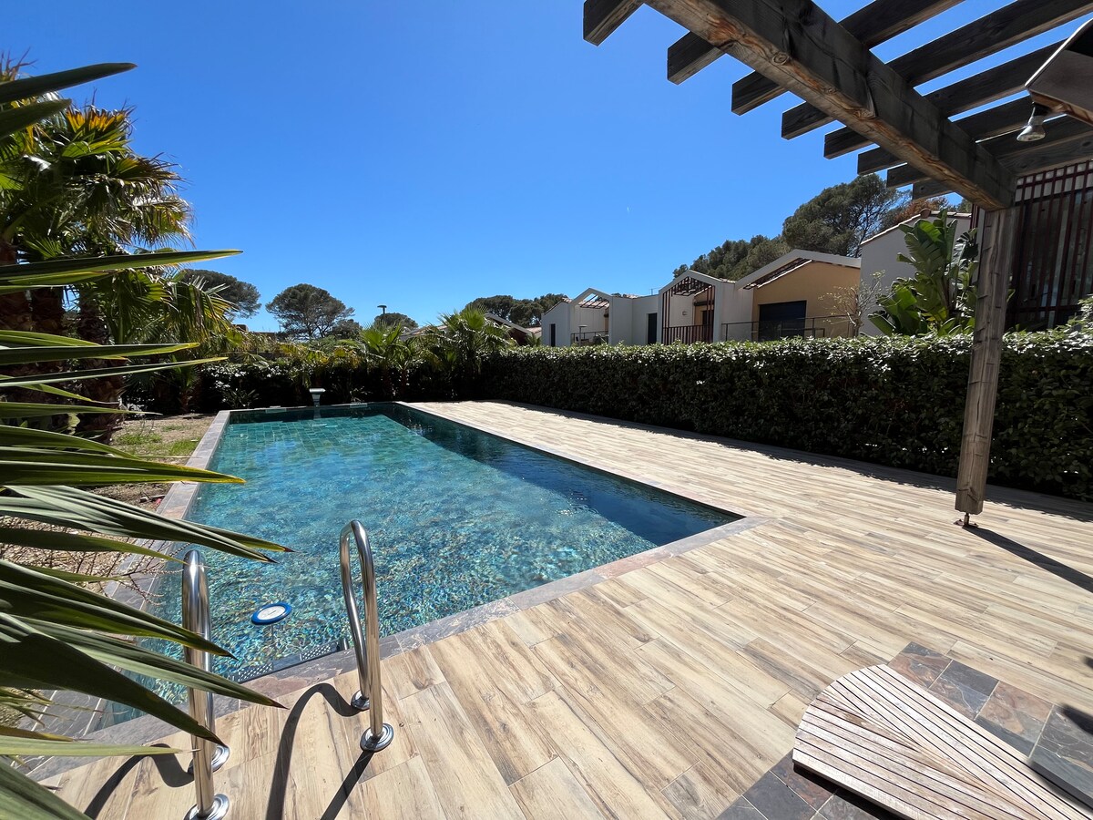 A refreshing swimming pool is framed by wooden decking and lush greenery, creating a relaxing outdoor space. Sunlight reflects off the clear water, inviting relaxation. Surrounding palm trees and planted hedges provide natural privacy, enhancing the tranquil atmosphere.