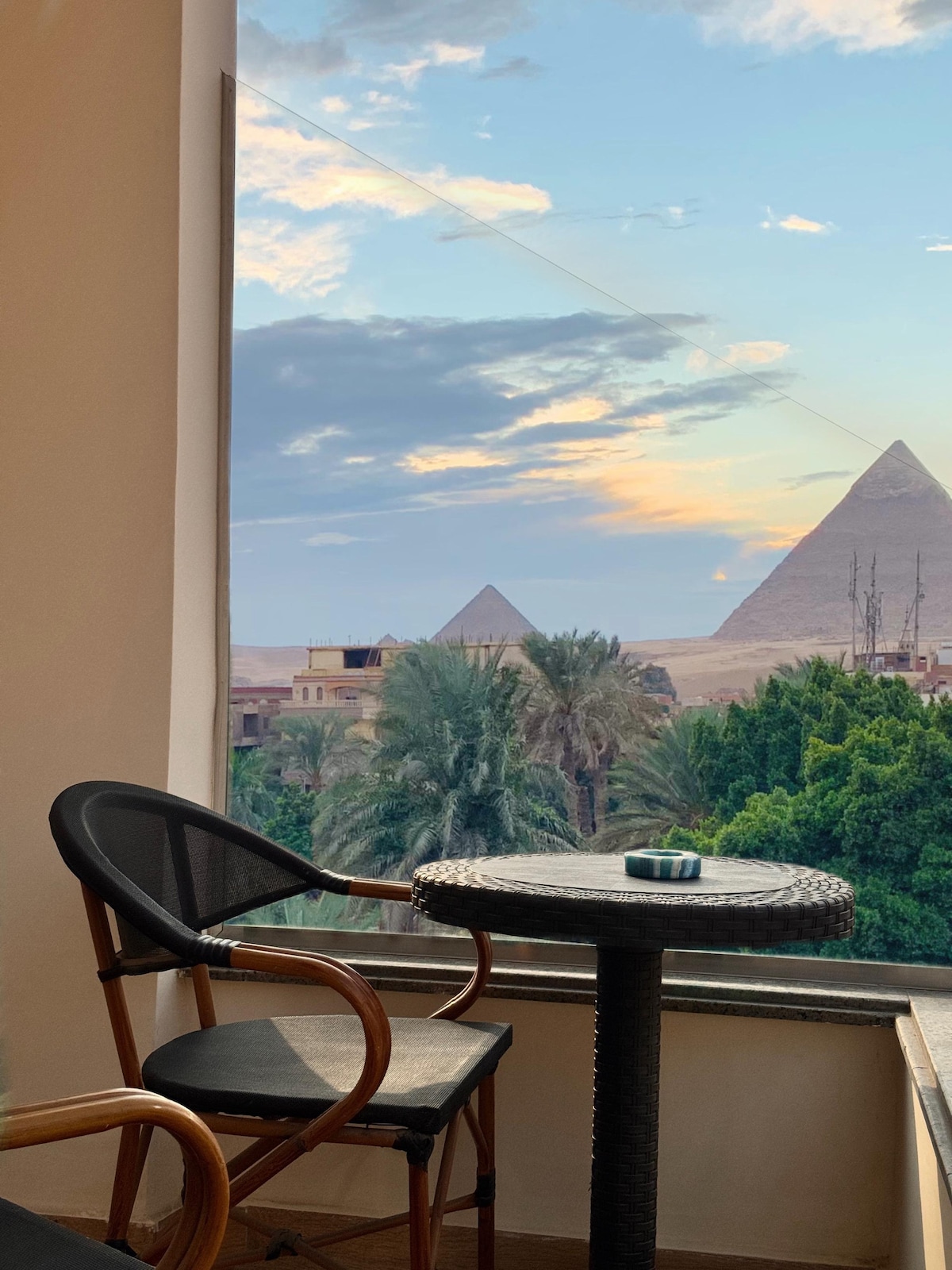 An outdoor terrace features a small round table and a chair, with a view of the pyramids framed by lush greenery and a colorful sky at dusk. The landscape combines natural and historical elements, creating a serene atmosphere.