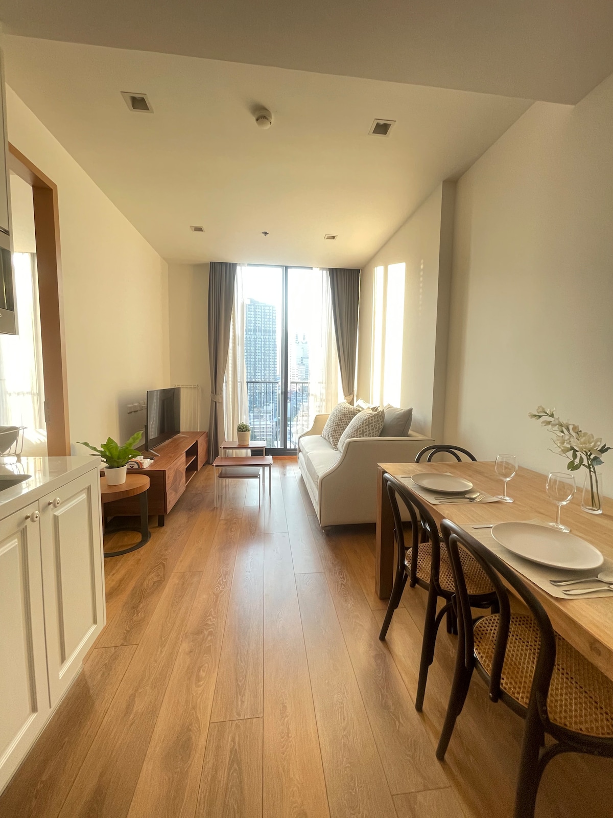 A bright living and dining area is showcased, featuring a comfortable sofa and a wooden dining table set for two. Large windows allow natural light to illuminate the space, highlighting the view of the city skyline. Minimalist decor enhances the sense of openness.