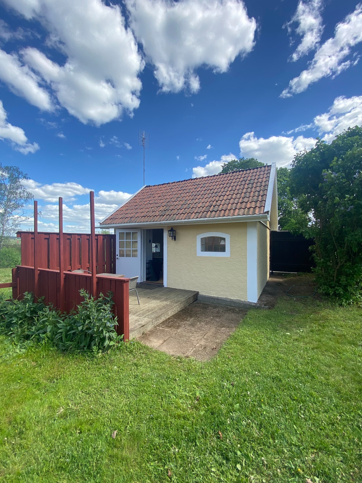 A small country house is depicted with a sloped red roof and light-colored walls. A wooden porch extends from the entrance, surrounded by green grass and plants. The clear blue sky features scattered clouds, enhancing the rural setting.