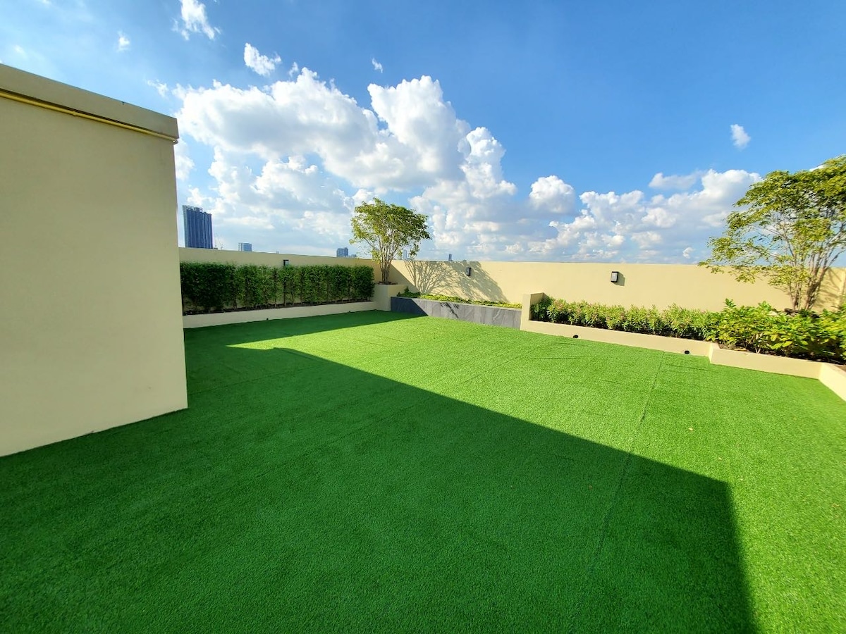 A spacious terrace is presented, featuring lush green artificial grass and bordered by neatly trimmed hedges. A few trees are visible in the background against a clear blue sky, creating an open and airy environment.