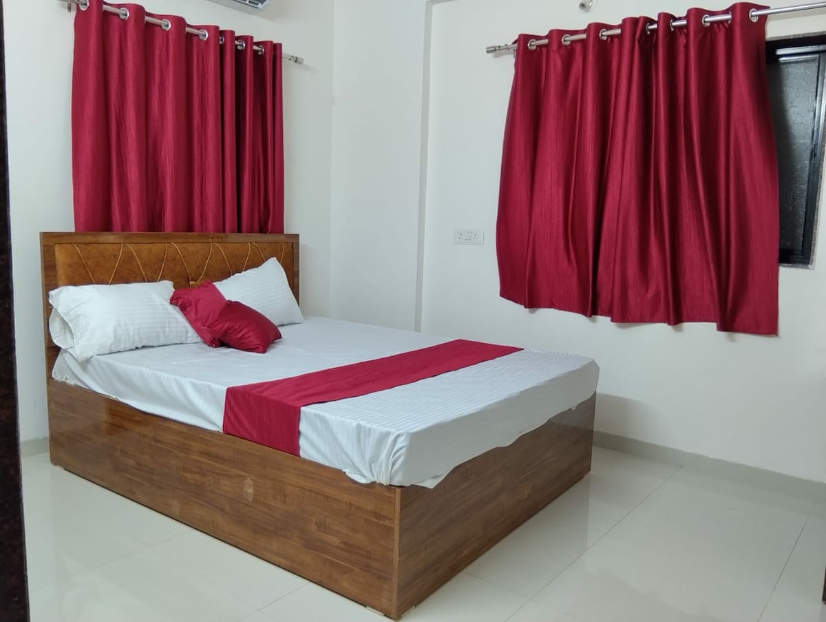 A spacious bed with a wooden frame is dressed in white linens, complemented by a red accent pillow. Red curtains frame a window, allowing natural light to fill the room, while light-colored tiles enhance the airy feel of the space.