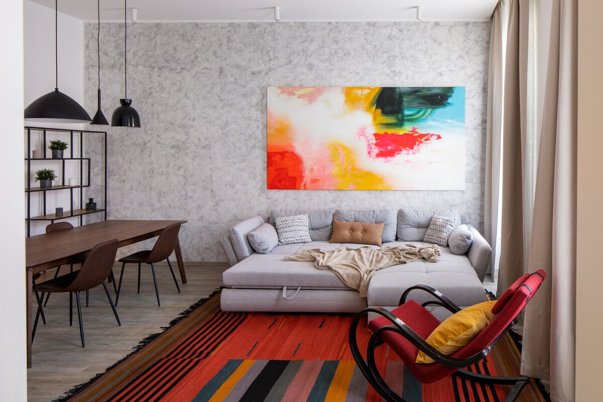 A modern living space features a gray sectional sofa adorned with a light-colored throw. A vibrant abstract painting hangs above the sofa, contrasting against the textured wall. A wooden dining table with several chairs is visible, accompanied by decorative plants on a shelving unit.