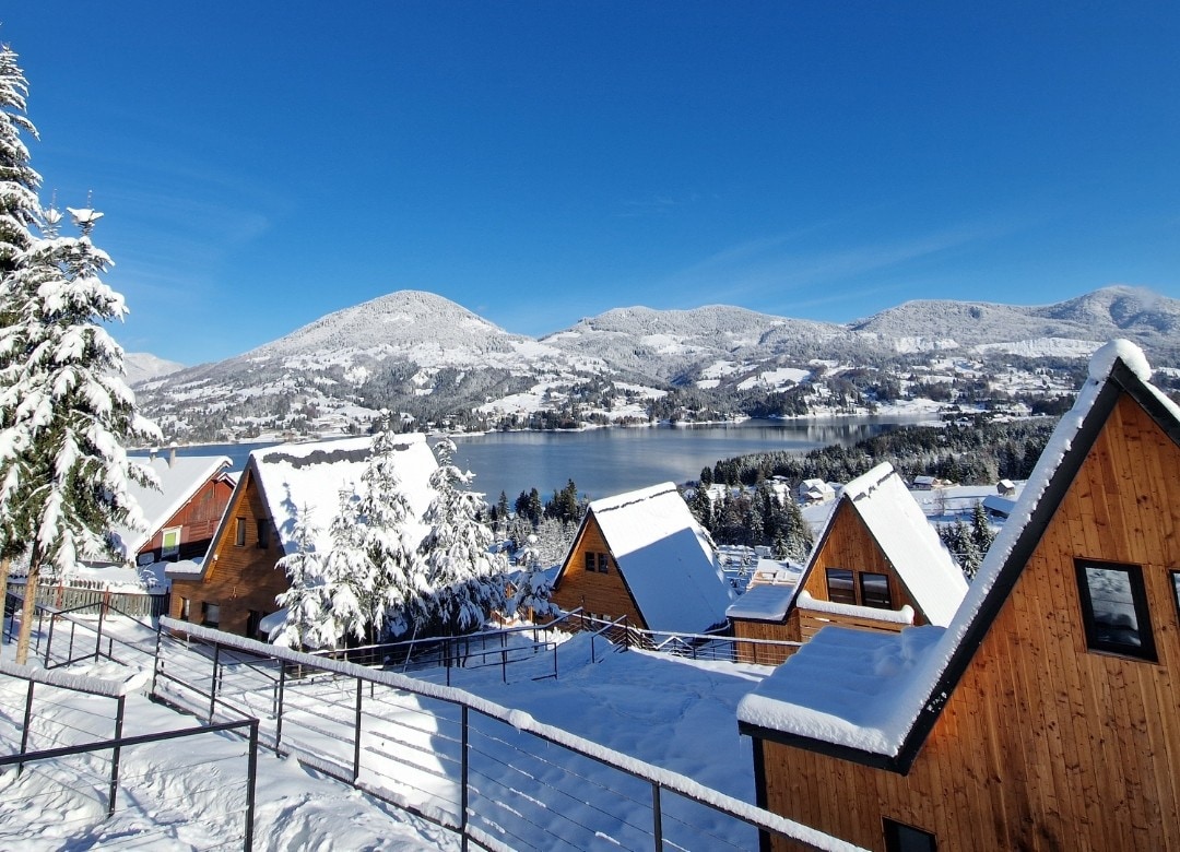 A snow-covered landscape features several A-frame cabins in the foreground, with varying roof angles. The serene lake and distant mountains provide a tranquil backdrop under a clear blue sky. The scene conveys a peaceful winter setting, inviting outdoor exploration and relaxation.